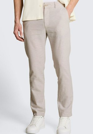 Mężczyzna w beżowych dopasowanych spodniach i białych trampkach, stojący z jedną ręką w kieszeni na jednolitym tle.