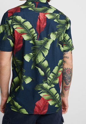 Mand iført mørkeblå kortærmet skjorte med stort grønt og rødt tropisk bladmønster, synlige tatoveringer på højre arm, set bagfra.