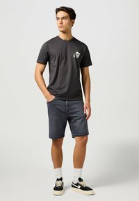 Camiseta de algodón gris oscuro con mangas cortas y un gráfico en el lado izquierdo del pecho, combinada con pantalones cortos de mezclilla oscura y zapatillas negras.