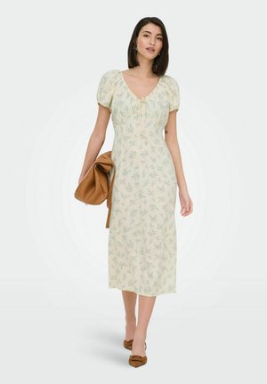 Femme portant une robe midi jaune pâle à fleurs avec des manches bouffantes, tenant une grande pochette marron pliée, et portant des chaussures pointues marron.