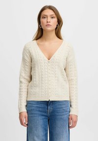 Krämfärgad stickad cardigan med V-hals, intrikat spetsmönster och pärlknappar, med ribbade ärmslut och nederkant. Buren över denim.