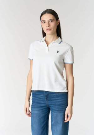 Giovane donna con lunghi capelli scuri che indossa una polo bianca con rifiniture blu e jeans blu, in piedi davanti a uno sfondo chiaro e uniforme.