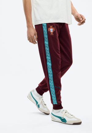 Persona che indossa pantaloni sportivi color granata con strisce laterali blu decorate e un emblema a scudo, abbinati a sneakers bianche e turchesi.