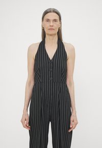 Svart stripet jumpsuit med halterneck, med en knappet front og en tettsittende midje, laget av et glatt stoff.