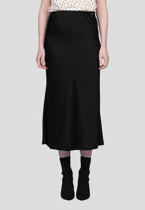 Jupe midi en satin noir avec une coupe droite, présentant une fente latérale et un accent logo discret à la taille. Portée avec des chaussettes noires.