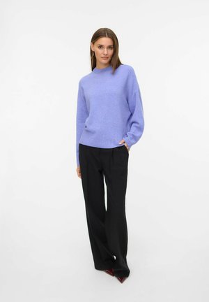 Pull en tricot lilas clair avec un col haut et un ourlet côtelé, associé à un pantalon noir à jambes larges et des chaussures rouges à bout pointu.