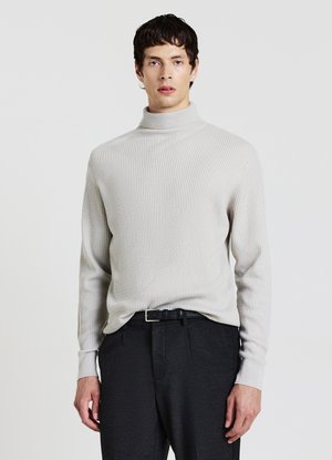 Giovane uomo che indossa un maglione dolcevita a coste grigio chiaro infilato in pantaloni grigio scuro con una cintura nera, in piedi davanti a uno sfondo semplice.