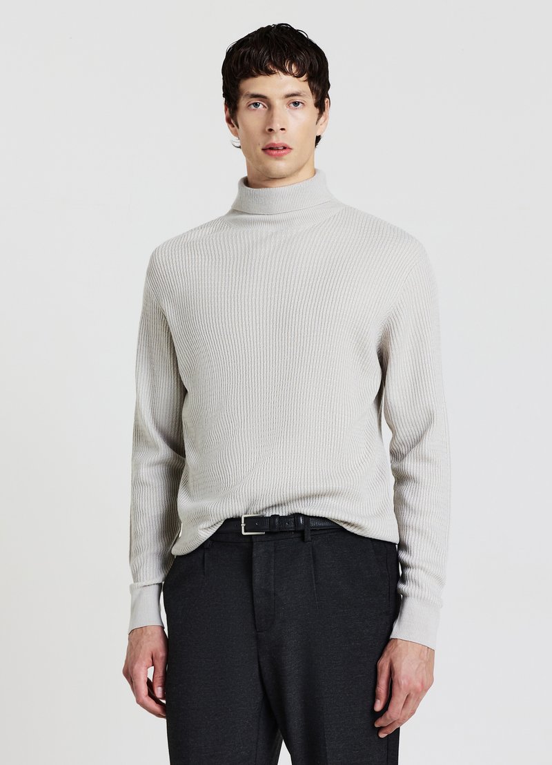 Jeune homme portant un pull à col roulé côtelé gris clair, rentré dans un pantalon gris foncé avec une ceinture noire, debout devant un fond uni.