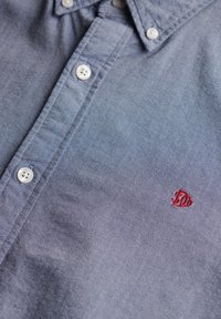 Niebieska koszula z guzikami w białym kolorze i małym czerwonym haftowanym logo na piersi, prezentująca detale kołnierza i listwy guzikowej.