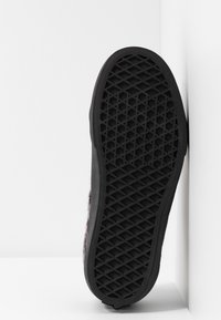 Schwarze Gummisohle eines Sneakers mit einem Waffelmuster, vertikal an einer weißen Ecke positioniert.