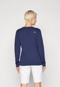 Pull en maille bleu marine à manches longues, col rond, avec des poignets et un ourlet côtelés, arborant un petit logo dans le haut du dos. Associé à un short blanc.