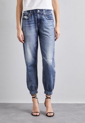 Femme portant un jean jogger en denim clair délavé avec des poignets élastiques, un détail usé sur une cuisse, associé à des sandales noires à talons hauts avec des brides.