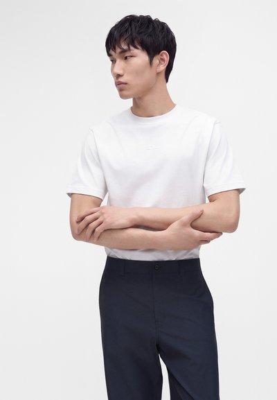 Jeune homme aux cheveux noirs courts, portant une chemise blanche unie à manches courtes et un pantalon foncé, debout bras croisés regardant vers la gauche.