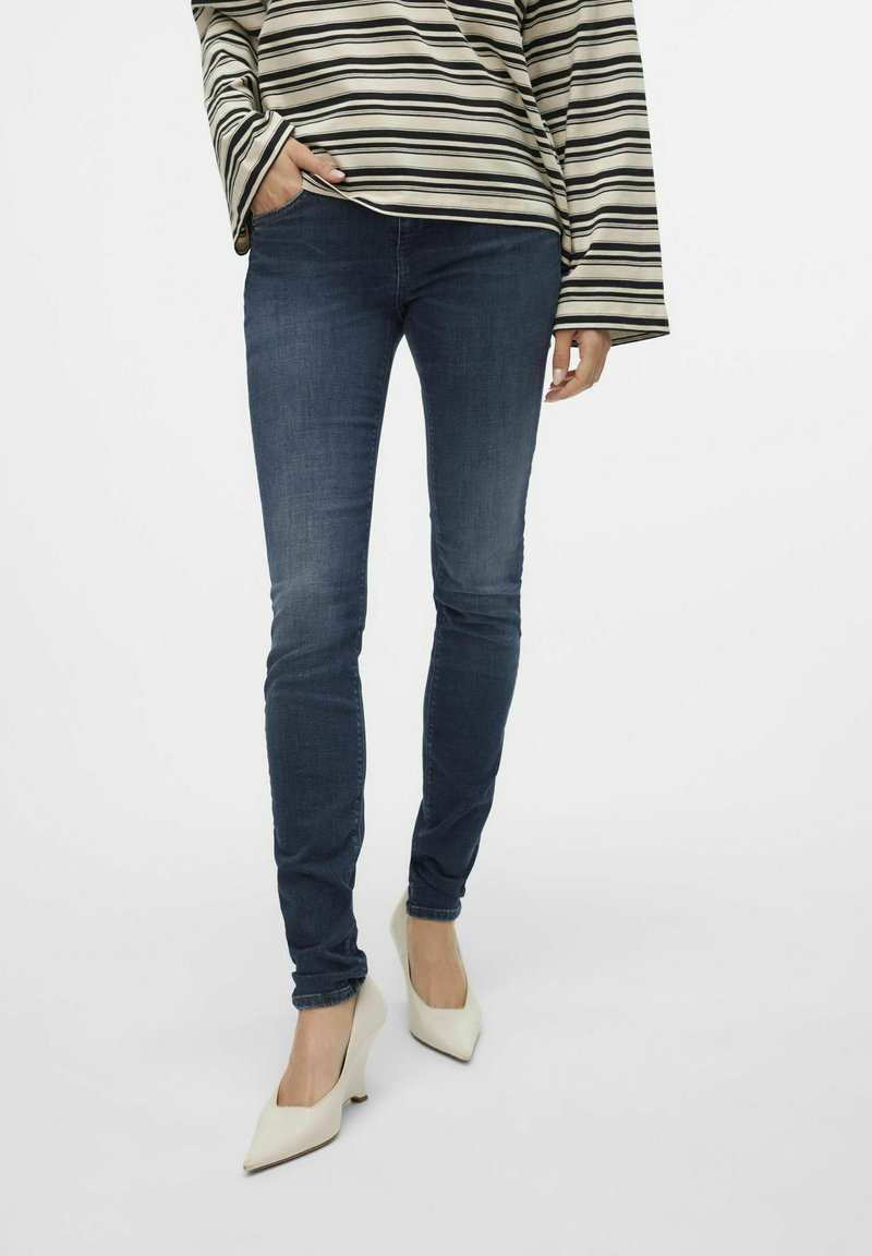 Tievie tumši zila džinsi no denima, apvienoti ar baltām izliekta papēža kurpēm. Augšdaļai ir horizontālas melnas un bēšas svītras ar platām piedurknēm.