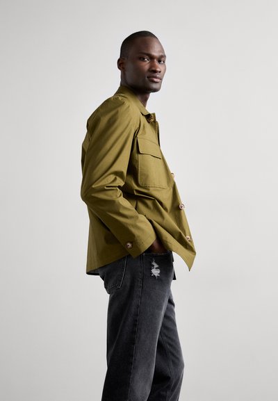 Olivenfarvet, skjorteblusejakke med to brystlommer, parret med slidte sorte jeans. Glat stof, afslappet pasform og enkelt design.