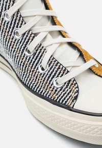 Detailní záběr na šněrovací tenisku s černobílým pruhovaným textilem, bílou podrážkou a žlutým lemováním okolo jazyka.