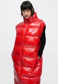 Piros puffermellény magas gallérral, fényes felülettel, túlméretezett dizájnnal, rövid ujjakkal és látható elülső cipzárral; két oldalzsebbel rendelkezik.