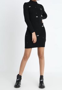 Robe noire côtelée à manches longues, col montant, ornée de nœuds et de perles dispersées. Associée à des bottines noires à chevilles.