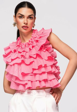 Kvinde med mørkt hår iført lagdelt, lyserødt rynket ærmeløst top og hvide bukser, poserer med den ene hånd på hoften mod grå baggrund.