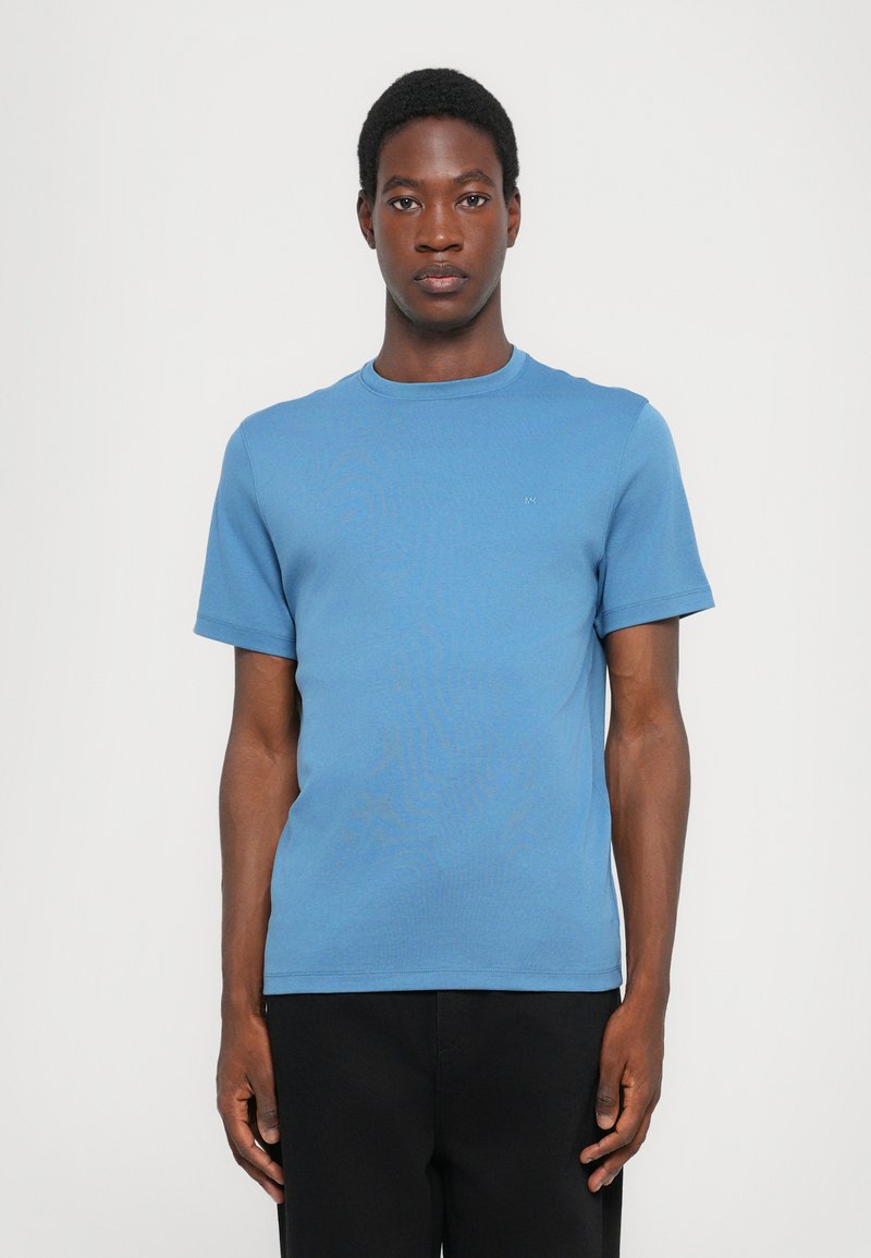 Svetlo modra majica s kratkimi rokavi iz mehkega materiala, z okroglim izrezom in majhnim logotipom na levem prsnem delu, v kombinaciji s črnimi hlačami.