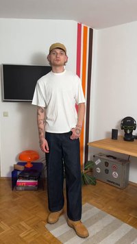 Hvit kortermet T-skjorte, mørke vide jeans, brune arbeidssko, og en lys brun caps. Tatoveringer synlige på armene. Enkelt bakgrunn.