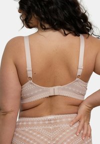 Soutien-gorge en dentelle beige avec bretelles réglables, présentant un motif géométrique et des bords festonnés. Fermeture au dos avec crochets et texture lisse.