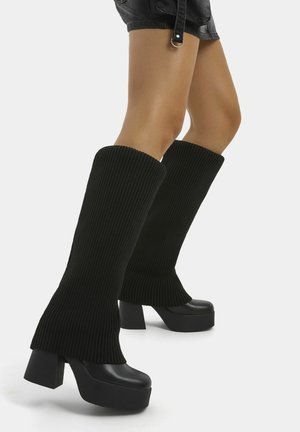 Jambes portant des chauffe-jambes en tricot côtelé noir par-dessus des bottes à talons compensés et un short en cuir noir sur un fond uni.