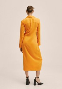 Helles orangefarbenes Kleid mit langen Ärmeln, einem Kragen und mittellanger Länge bis zur Wade. Glatte Textur; kombiniert mit schwarzen Schuhen mit Blockabsatz.