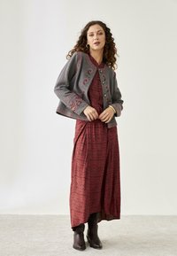 Chaqueta gris corta con bordados florales, combinada con un vestido largo rojo de estampado y botas de tobillo marrones. Tela texturizada y detalles visibles.