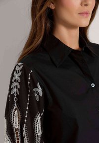 Schwarzes Hemd mit Kragen und Knopfverschluss; verfügt über weiße bestickte Blumenmuster an den Ärmeln und hat eine glatte Textur.
