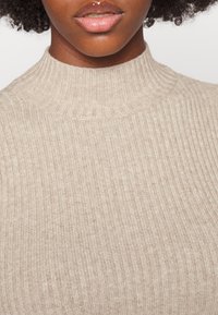Maglione girocollo a coste beige con colletto alto e texture morbida. Vista ravvicinata che mostra il fine dettaglio del lavoro a maglia e una distribuzione uniforme del colore.