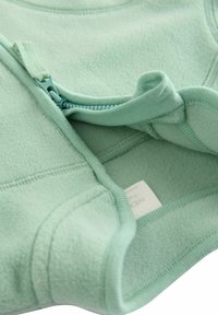 Hanorac din polar verde mentă, cu un închidere prin fermoar, textură moale și margini cu elastic subțire. Eticheta este vizibilă în interior.