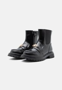 Bottes de cheville noires brillantes avec semelles épaisses, zips latéraux, panneaux élastiques et détails en chaîne dorée sur les sangles avant, sur fond blanc.