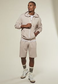 Mann in beigem Reißverschluss-Hoodie und passender Shorts, darunter ein weißes T-Shirt, gestreifte Socken und weiße Sneaker, steht vor einem schlichten Hintergrund.