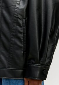Schwarze Lederjacke mit glatter Textur, ausgestattet mit einer Seitentasche und einem knöpfbaren Manschettendetail, das ein übergroßes Design präsentiert.
