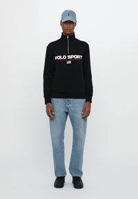 Juodas zipas su užtrauktuku su "POLO SPORT" logotipu, šviesiai mėlynos džinsinės kelnės ir juodi batai. Aksesuarai apima šviesiai mėlyną kepurę.