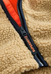 béžová fleecová látka s texturovaným povrchem, uprostřed černý zip lemovaný jasně oranžovou barvou a štítek s nápisem "SUPERDRY."