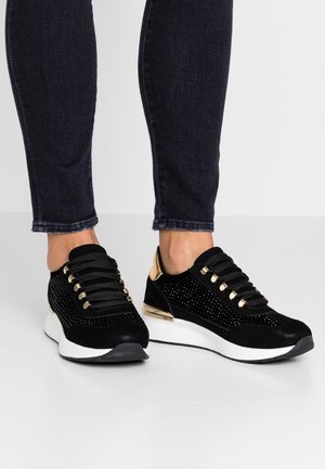 Czarne sneakersy ze złotymi akcentami i czarnymi sznurówkami, noszone z ciemnymi obcisłymi dżinsami, stojące na jasnoszarej podłodze i na białym tle.