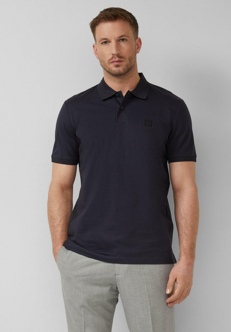 Homme portant un polo bleu marine à manches courtes avec col et un pantalon gris clair, debout devant un fond uni.