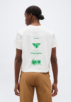 Frau mit geflochtenem Haar, die ein weißes, kurzes T-Shirt mit grünem "Gletscher schmilzt"-Aufdruck und braune Hose trägt, von hinten gesehen.