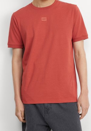 Camiseta de algodón roja con cuello redondo y mangas cortas, que presenta un pequeño logotipo cuadrado en el centro. Textura suave y corte relajado.