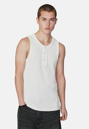 Junger Mann mit kurzen Haaren, der ein weißes ärmelloses Hemd mit Knöpfen und dunkle Hosen trägt, vor einem schlichten hellen Hintergrund stehend.