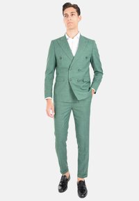 Abito verde doppiopetto in tessuto testurizzato, pantaloni slim-fit e camicia bianca. Presenta mocassini neri e una silhouette moderna.