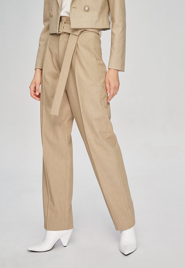 Beige op maat gemaakt pak met een cropped jasje, wijde broekspijpen en een ceintuur om de taille. Witte puntige enkellaarzen maken de look compleet.