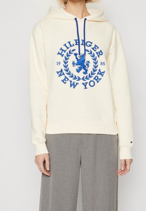 Osoba nosząca kremową bluzę z kapturem z niebieskim napisem „Hilfiger New York 1985” i herbem lwa, w połączeniu z szarymi spodniami.