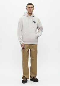 Light beige hooded sweatshirt with a front kangaroo pocket and navy logo, paired with khaki trousers and dark shoes on a plain background.
