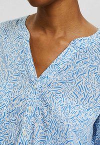 Blaue und weiße gemusterte Bluse aus leichtem Stoff mit V-Ausschnitt, Knopfleiste und strukturiertem Design mit wellenförmigen Linien.