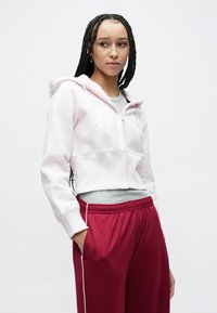 Ružová krátka mikina na zips s kapucňou, v kombinácii s voľnými červenými nohavicami s bielymi bočnými pruhmi a elastickým pásom.