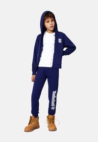 Un sweat à capuche zippé bleu marine sur un t-shirt blanc, assorti à un pantalon de survêtement bleu marine portant le logo "Timberland", complété par des bottes beiges.