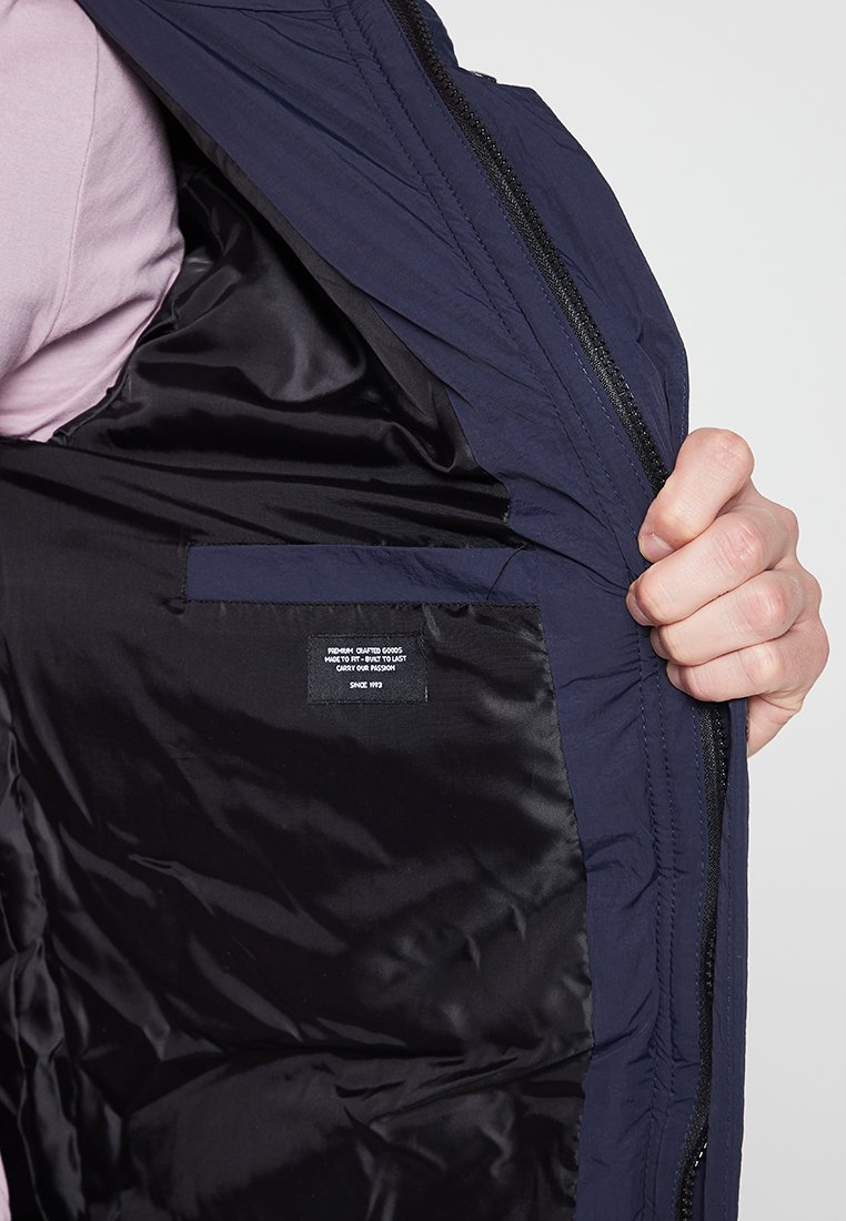 Veste isolée bleu marine avec une doublure intérieure lisse noire, dotée d'une fermeture éclair et d'une poche intérieure avec une étiquette.